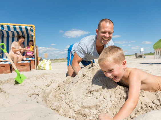 Lachende vader en zoon bouwen samen een groot zandkasteel op het strand, met een klassieke Duitse strandstoel op de achtergrond.