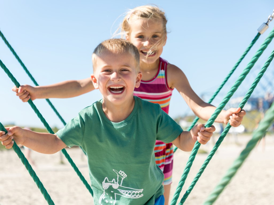 Lachende broer en zus klimmen samen in een groen touwenparcours op het strand.