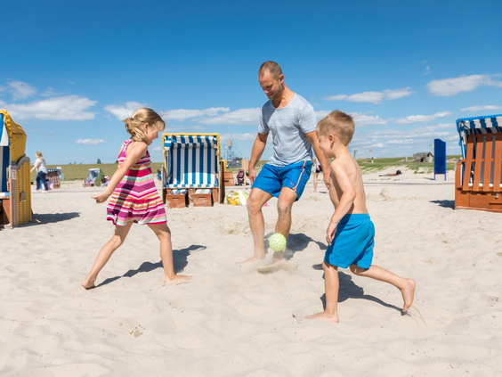 Vader speelt een potje voetbal met zijn zoon en dochter op het zandstrand voor de blauw-wit gestreepte strandstoelen.