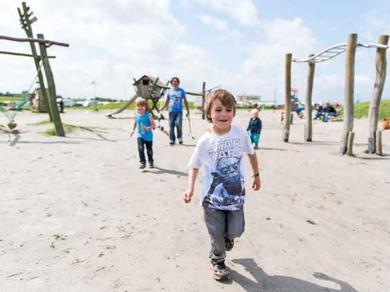 Jongen rent vrolijk door een avontuurlijke strandspeeltuin met houten speeltoestellen aan de Noordzeekust.