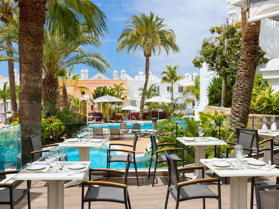 Gasten genieten van hun maaltijd aan tafels op het buitenterras van het restaurant, onder een pergola en met uitzicht op de tropische tuin.