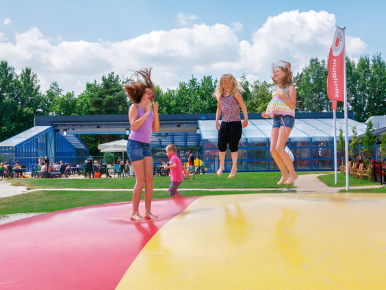 Kinderen springen en lachen op de grote, kleurrijke airtrampoline in de buitenspeeltuin van Landal het Land van Bartje, met het zwembad op de achtergrond.
