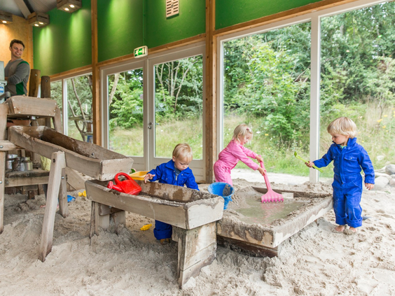 Kinderen in overalls spelen in de binnenspeeltuin Bartje's Boerderij, met een grote zandbak, houten speeltoestellen en uitzicht op het bos.
