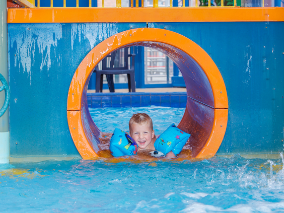 Een lachende jongen met zwemvleugels kijkt door een opening in de oranje tunnel van een waterglijbaan in het zwembad van Landal het Land van Bartje