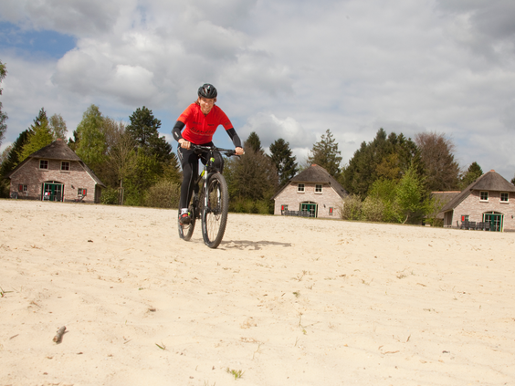 Een jongen op een mountainbike fietst over een zandvlakte op het vakantiepark Landal het Land van Bartje, met Drentse boerderijen op de achtergrond.