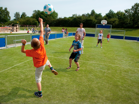 Een groep kinderen en volwassenen speelt een partijtje voetbal op het omheinde kunstgras sportveld van Landal het Land van Bartje.
