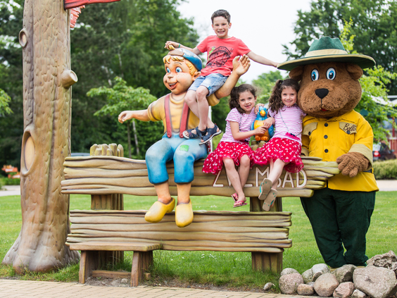 Een groep lachende kinderen poseert voor een foto op een bankje met de mascottes Bartje en Bollo de Beer op vakantiepark Landal het Land van Bartje.