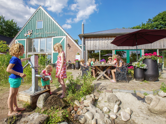 Kinderen spelen met de waterpomp en waterbaan op het terras bij Bartje's Boerderij op Landal het Land van Bartje, terwijl gasten op de achtergrond zitten.