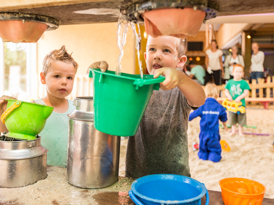 Twee jonge kinderen spelen enthousiast met emmers, zand en een zandlift in de overdekte zandspeelplaats van Bartje's Boerderij op Landal het Land van Bartje.