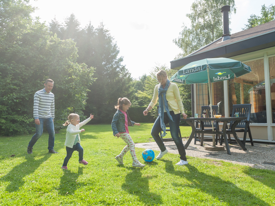 Een gezin met twee kinderen speelt vrolijk een potje voetbal in de tuin bij hun vakantiebungalow op Landal Aelderholt, met een terras en parasol op de achtergrond.