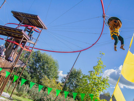 Een kind met een gele helm en veiligheidsharnas zoeft door de lucht aan de rode skyflyerl van het klimpark op Landal Aelderholt, met een blauwe lucht op de achtergrond.
