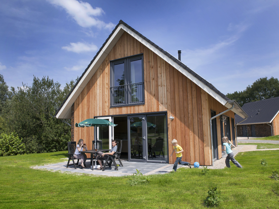 Een moderne, duurzame houten bungalow op Landal Aelderholt, waar ouders op het terras zitten en kinderen op het grasveld ervoor spelen op een zonnige dag.