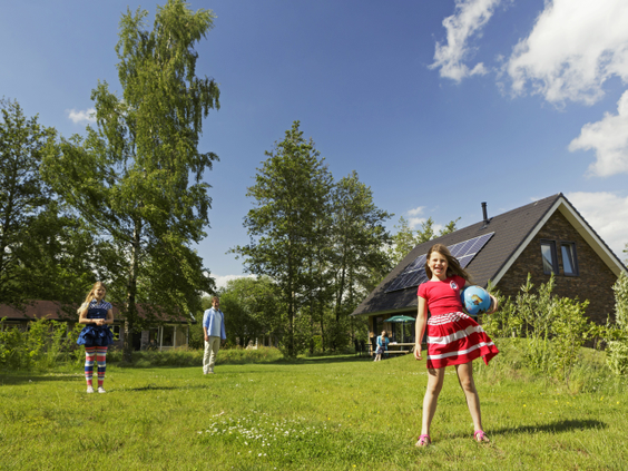 Drie kinderen spelen samen met een bal op een groot grasveld voor hun vakantiebungalow op een zonnige dag bij Landal Aelderholt.