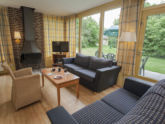 Een knus ingerichte woonkamer met een comfortabele zithoek, open haard en grote ramen met uitzicht op het groene buitengedeelte van de bungalow op Landal Aelderholt.