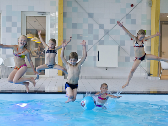 Een groep enthousiaste kinderen springt tegelijk met een grote plons in het blauwe water van het binnenzwembad op Landal Amerongse Berg.