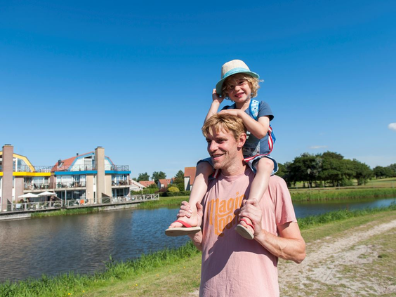 Een vader wandelt met zijn kind op de schouders langs het water op Landal Beach Resort Ooghdyne, met het centrumgebouw op de achtergrond.