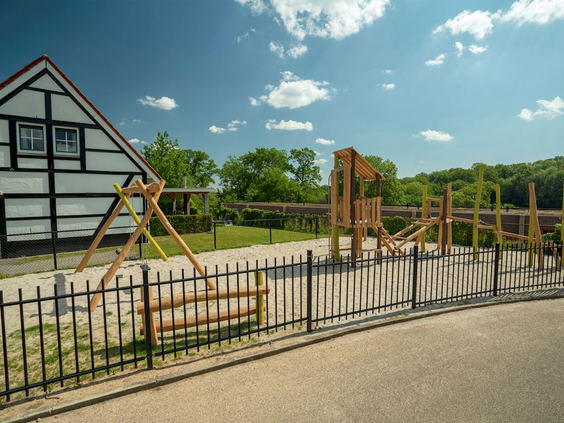 Uitzicht vanaf een pad op de houten speeltuin van vakantiepark Landal Cauberg, met de kenmerkende vakwerkhuizen op de achtergrond.