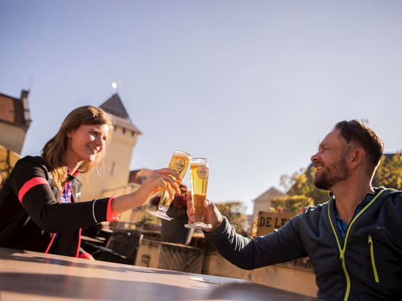 Een stel proost met een drankje op een zonnig terras in Valkenburg, met op de achtergrond de Kasteelruïne.