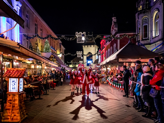 Dansende figuren in rode pakjes tijdens de kerstparade in een sfeervol verlichte straat met volle terrassen in Valkenburg.