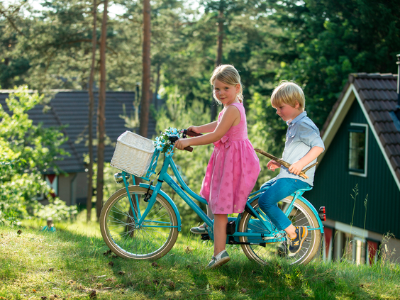 Een jongen en een meisje rijden samen achterop een blauwe fiets over een grasveld op vakantiepark Landal Coldenhove.
