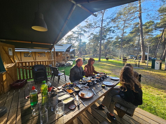 Een gezin eet gezellig buiten op de houten veranda van een safaritent op Landal Coldenhove, midden in het bos.