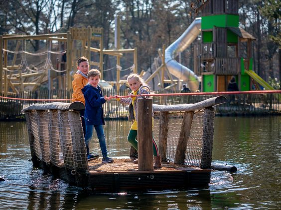 Kinderen op een houten trekvlot in de speelvijver met op de achtergrond de grote avontuurlijke buitenspeeltuin van Landal De Lommerbergen in Limburg.