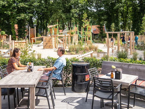 Ouders op het terras van het restaurant met direct uitzicht op de kinderen die spelen in de natuurspeeltuin van Landal De Vers.
