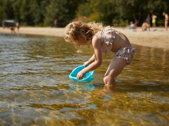 Klein meisje met krullen speelt met een schepje in het ondiepe water van de recreatieplas bij Landal De Wedderplassen.