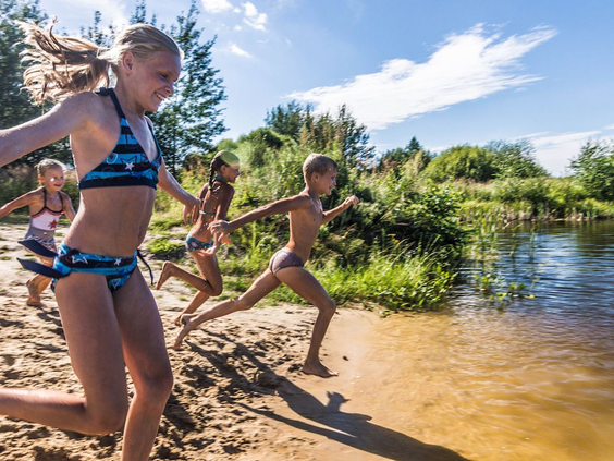 Een groep kinderen rent lachend vanaf een zandstrandje een recreatiemeer in op vakantiepark Landal Drentse Lagune in Drenthe.