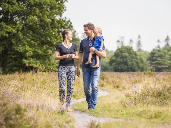 Een jong gezin wandelt over een zandpad door een Drents heidegebied, omringd door paarse heide en groene bossen.