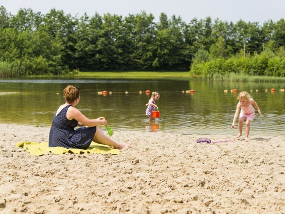 Een moeder kijkt toe vanaf een handdoek op het zandstrand terwijl haar jonge kinderen spelen in het ondiepe water van de recreatieplas bij Landal Drentse Lagune.