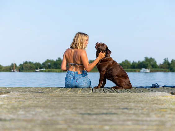 Een vrouw met haar bruine hond geniet van het uitzicht op een houten vlonder aan de rand van een recreatieplas in Drenthe.