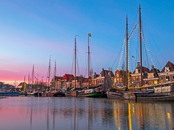 Een haven vol met traditionele Friese zeilschepen (skûtsjes) die weerspiegelen in het water tijdens een kleurrijke zonsondergang.