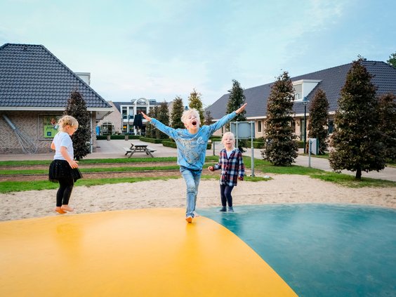 Drie kinderen springen en balanceren met hun armen wijd op het grote, kleurrijke luchtkussen in de buitenspeeltuin.