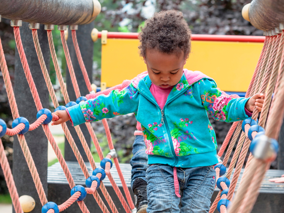 Een peuter in een kleurrijk vest balanceert voorzichtig over een wiebelende touwbrug in de buitenspeeltuin van het vakantiepark.