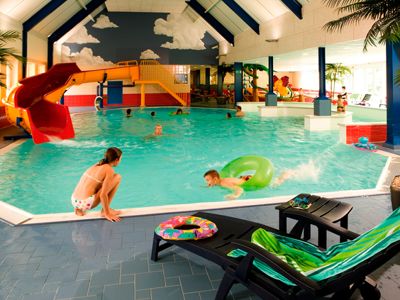 Het overdekte zwembad van Landal Hellendoornse Berg met een glijbaan, zwemmende kinderen en ligbedden langs de kant.