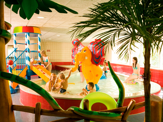 Kinderen spelen vrolijk in het kinder-doe-bad, compleet met een spuitende olifant en speeltoestellen onder een palmboom.