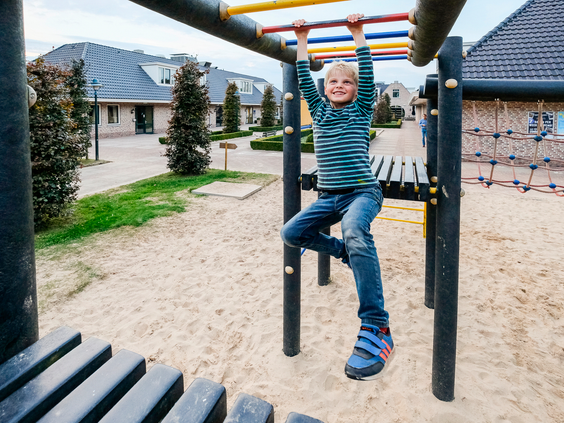 Een stoere jongen hangt aan het klimrek in de buitenspeeltuin van Landal Hellendoornse Berg, omringd door zand en andere speeltoestellen.