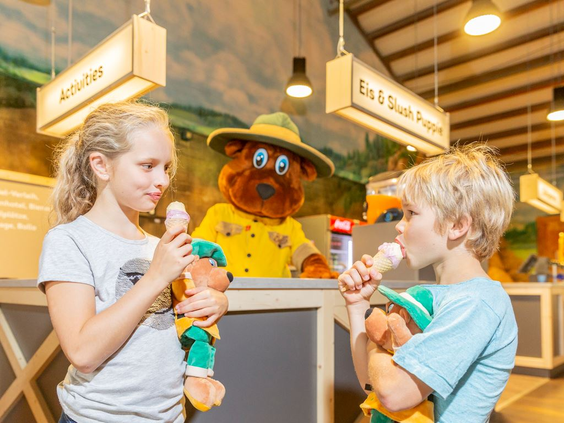 Twee kinderen eten een ijsje dat ze hebben gekregen van mascotte Bollo de Beer in het centrumgebouw van Landal Hochwald.