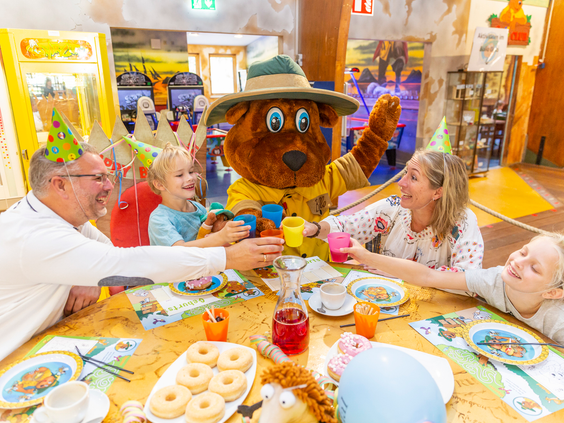 Een familie met kinderen viert een verjaardagsfeestje met mascotte Bollo en proost met feestmutsen op bij Landal Hochwald.