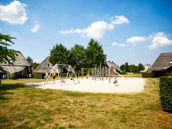 Een breed overzicht van een grote zandspeeltuin met diverse houten speeltoestellen, schommels en een glijbaan, gelegen naast de Saksische boerderijen van Landal Hof van Saksen.