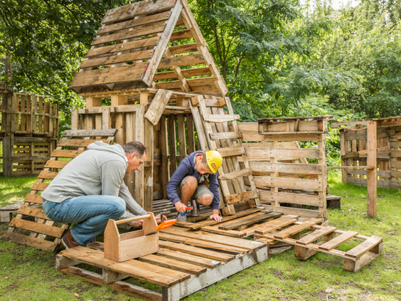 Een vader en zoon zijn samen een hut aan het bouwen van houten pallets en ander bouwmateriaal tijdens de kinderactiviteit hutten bouwen bij Landal Hof van Saksen.