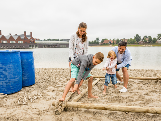 Een gezin werkt samen op het strand om met tonnen en houten palen een eigen vlot te bouwen, een avontuurlijke workshop bij Landal Hof van Saksen.