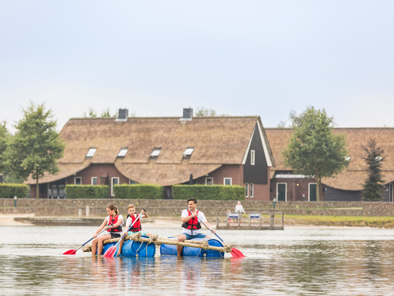 Drie kinderen met zwemvesten aan peddelen op hun zelfgebouwde vlot van blauwe tonnen op het recreatiemeer van Landal Hof van Saksen.