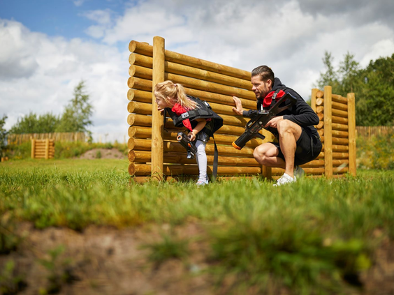 Twee kinderen verstoppen zich achter een houten obstakel in de outdoor lasergame-arena van Landal Hof van Saksen, terwijl ze proberen hun tegenstanders te raken.