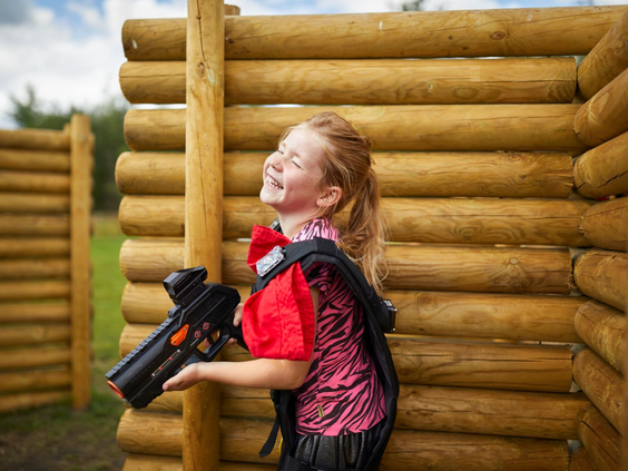Een lachend meisje met een lasergun en een vest dat rood oplicht, leunt tegen een houten muur in de outdoor lasergame-arena van Landal Hof van Saksen.