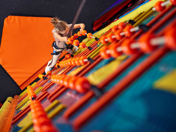 Een jongen, gezekerd met een tuigje, beklimt een unieke en kleurrijke klimwand met oranje en gele sporten in de indoor speelhal van Landal Hof van Saksen.