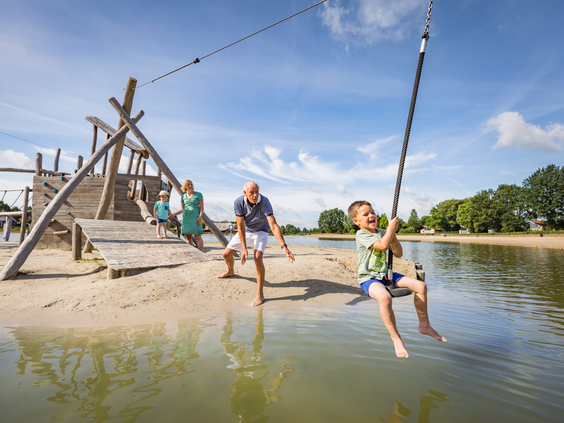 Een jongen zoeft met de kabelbaan over het water van het recreatiemeer bij Landal Hof van Saksen, terwijl zijn vader vanaf het strandje toekijkt.