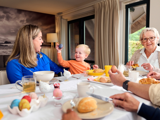 Een familie met opa, oma, ouders en een klein kind zit aan een feestelijk gedekte ontbijttafel met gekleurde paaseieren in hun vakantieboerderij op Landal Hof van Saksen.