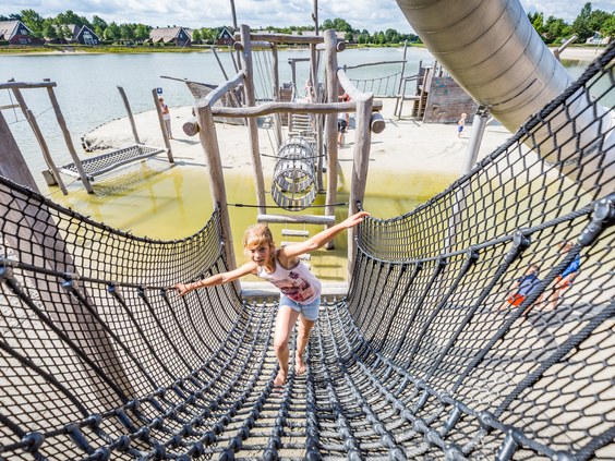 Een meisje balanceert met haar armen wijd op een lange touwbrug die onderdeel is van het grote houten speeltoestel bij het recreatiemeer van Landal Hof van Saksen.